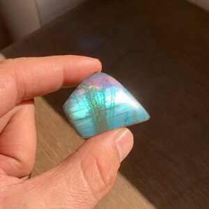 May include: A polished, triangular labradorite gemstone. The stone displays a vibrant play of color, with iridescent flashes of blue, green, and purple across a base of gray and blue. The gemstone is held in a hand.