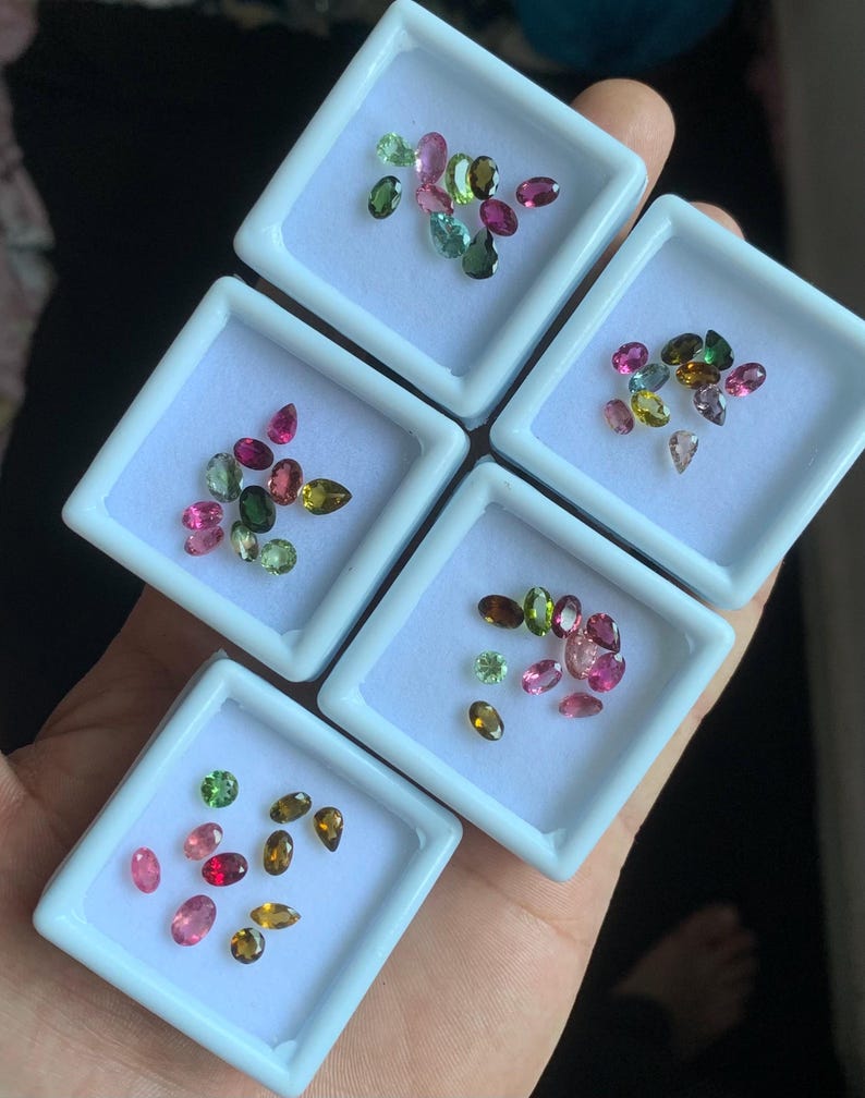 Puede incluir: Cinco bandejas cuadradas blancas contienen cada una una colecci&oacute;n de gemas de colores. Las gemas son ovaladas y redondas, en tonos de rosa, verde, marr&oacute;n y amarillo. Las bandejas se sostienen en una mano.