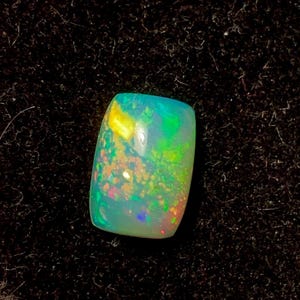 Puede incluir: Una piedra preciosa de ópalo rectangular con esquinas redondeadas. El ópalo muestra un vibrante juego de colores, con destellos de verde, azul, amarillo y naranja. La piedra está sobre un fondo oscuro y texturizado, resaltando sus cualidades iridiscentes.
