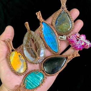 May include: A collection of handcrafted gemstone pendants in various shapes and colors, including blue, yellow, green, and pink. Each stone is intricately wrapped in copper wire, showcasing unique textures and patterns. The pendants are displayed against a dark background.