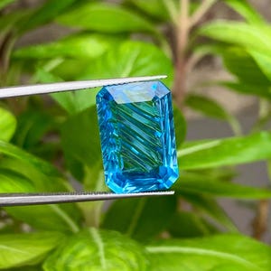 Könnte beinhalten: Ein rechteckiger, facettierter, leuchtend blauer Edelstein wird von einer Pinzette vor einem Hintergrund aus grünen Blättern gehalten. Der Edelstein hat ein lebendiges, transparentes Aussehen mit inneren linearen Mustern, die seinen Schliff und seine Klarheit zeigen. Die Farbe des Edelsteins ist ein lebendiges, klares Blau.