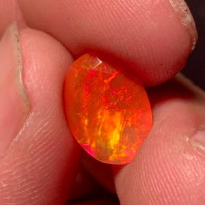 Puede incluir: Una vibrante piedra preciosa de ópalo facetada, que muestra una ardiente mezcla de tonos naranja, rojo y amarillo. La estructura interna de la gema crea un juego de luces, realzando su brillo. El ópalo se sostiene entre los dedos, resaltando su tamaño y color.