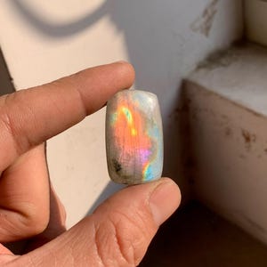 Könnte beinhalten: Ein rechteckiger, polierter Edelstein mit Regenbogen-Irisieren. Der Stein zeigt ein Farbspiel, darunter Orange, Rosa und Blau, vor einem grauen Hintergrund. Der Edelstein wird zwischen zwei Fingern gehalten.