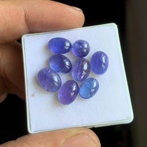 May include: A collection of oval-shaped, blue-violet gemstones displayed on a white surface. The gemstones vary slightly in color, with some appearing more purple and others more blue. The stones are smooth and polished, reflecting light.
