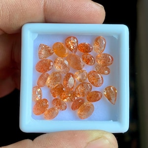Puede incluir: Una colección de piedras preciosas de piedra solar naranja en un recipiente cuadrado blanco. Las piedras preciosas tienen forma ovalada y de lágrima, con una apariencia brillante y translúcida. Las piedras son de color naranja cálido.