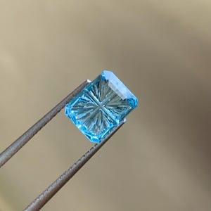 Könnte beinhalten: Ein quadratischer, hellblauer Edelstein mit einem Sternmuster wird von einer Metallpinzette gehalten. Der Edelstein hat eine facettierte Oberfläche und eine helle, klare Farbe. Der Hintergrund ist ein weicher, neutraler Ton.