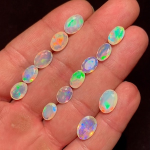 May include: A collection of oval-cut opals, each displaying a unique play of color with flashes of green, orange, and blue. The gemstones are held in a hand against a dark background, showcasing their iridescent qualities.