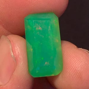 Könnte beinhalten: Ein lebendiger, rechteckig geschliffener Edelstein mit einem leuchtenden Grünton. Der Edelstein hat eine facettierte Oberfläche, die das Licht reflektiert und einen funkelnden Effekt erzeugt. Der Stein wird zwischen Fingern gehalten, was seine Klarheit und Farbe hervorhebt.