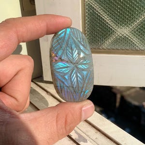 May include: An oval-shaped, iridescent labradorite gemstone with a carved leaf pattern. The stone displays shades of blue, green, and purple. The gemstone is held between fingers, showcasing its polished surface and intricate design.