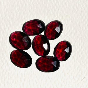 Könnte beinhalten: Sieben facettierte ovale Edelsteine in einem tiefen Rot. Die Edelsteine sind in einer Gruppe auf einer weißen Oberfläche angeordnet.