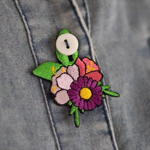May include: A close-up of a denim jacket with a floral embroidered patch. The patch features pink, purple, and yellow flowers with green leaves. A white button is visible on the jacket.