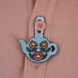 Puede incluir: Un suéter rosa con un botón bordado de una tetera azul. La tetera tiene flores rojas con un centro amarillo.