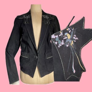 Può includere: Blazer nero con cuciture bianche e chiusura a un bottone. La giacca è esposta su un manichino. Viene mostrato anche un pezzo decorativo abbinato con ricami di uccelli e fiori. Lo sfondo è di colore rosa.