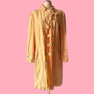 May include: A light orange trench coat with a unique button closure down the front. The coat has long sleeves and a collar. The coat is displayed on a mannequin against a pink background.