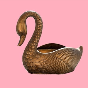 Könnte beinhalten: Eine metallisch bronzefarbene Schale in Schwanenform mit detaillierten Federstrukturen entlang des Halses und der Flügel. Der Schwan hat einen gebogenen Hals und eine hohle Mitte mit glatter Innenoberfläche und einer realistischen Optik.