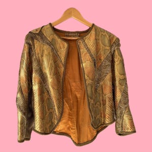 Könnte beinhalten: Eine Vintage-Jacke mit Schlangenmuster in Gold- und Brauntönen hängt an einem Holzbügel vor einem rosa Hintergrund. Die Jacke hat ein einzigartiges Design mit einem kragenlosen Ausschnitt und einer taillierten Passform, die eine luxuriöse Ästhetik zeigt.