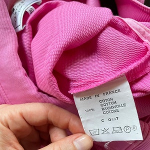 Może przedstawiać: Zbliżenie na jaskrawor&oacute;żowy element odzieży o teksturowym splocie. Etykieta głosi "MADE IN FRANCE" i wyszczeg&oacute;lnia skład materiału jako 100% bawełny. Etykieta zawiera r&oacute;wnież instrukcje prania i numer "40".