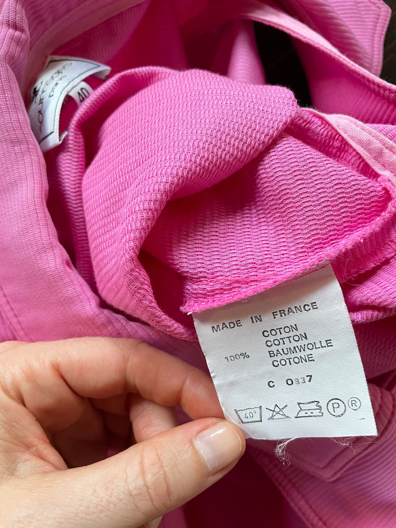 Może przedstawiać: Zbliżenie na jaskrawor&oacute;żowy element odzieży o teksturowym splocie. Etykieta głosi "MADE IN FRANCE" i wyszczeg&oacute;lnia skład materiału jako 100% bawełny. Etykieta zawiera r&oacute;wnież instrukcje prania i numer "40".