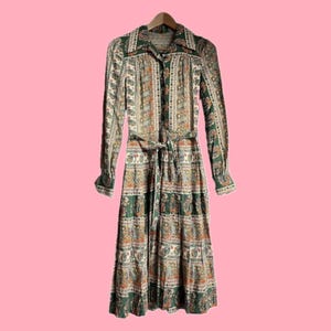 Può includere: Un abito vintage a maniche lunghe con abbottonatura e cintura annodata. L'abito presenta un colorato motivo floreale nei toni del verde, arancione e bianco. L'abito è appeso a una gruccia di legno su sfondo rosa.