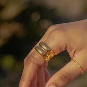 Könnte beinhalten: Nahaufnahme einer Hand, die zwei goldene Ringe am Ringfinger und einen dünnen goldenen Ring am Daumen trägt.