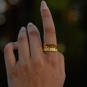 Könnte beinhalten: Ein goldener Hammerring an einem Finger. Der Ring ist breit und hat eine strukturierte Oberfläche.