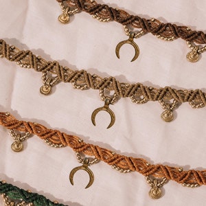 Può includere: Tre collane sovrapposte con cordoncino marrone e verde, perline dorate e ciondoli a forma di mezzaluna.