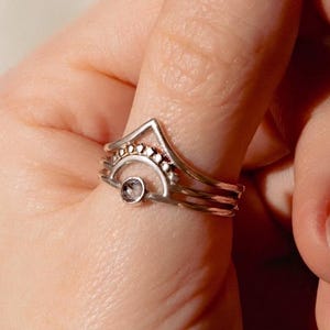Könnte beinhalten: Ein silberner Ring mit einem kleinen, runden, klaren Edelstein und einer Reihe kleiner, klarer Edelsteine. Der Ring hat ein mehrschichtiges Design mit einer dreieckigen Spitze. Der Ring befindet sich an einem Finger.