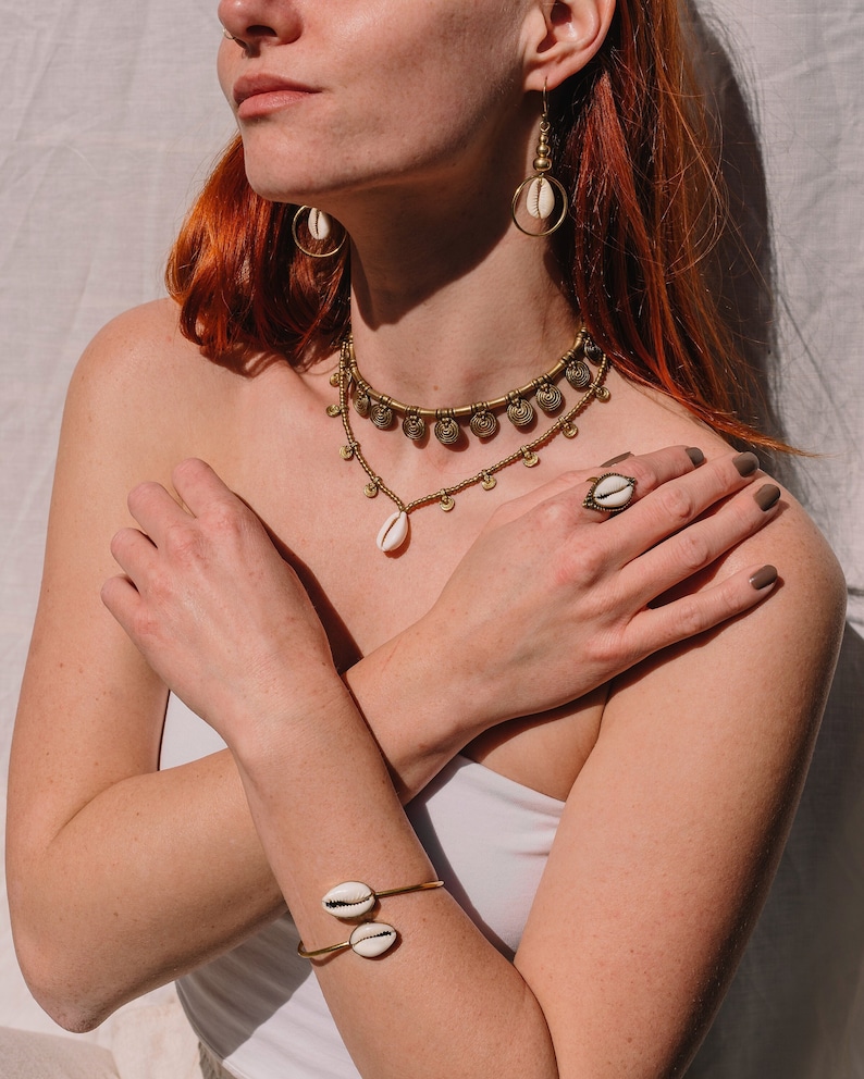 Puede incluir: Un conjunto de joyas doradas con conchas de cauri. El conjunto incluye un collar, pendientes, un anillo y una pulsera. El collar tiene m&uacute;ltiples filas con peque&ntilde;os dijes en forma de moneda. El anillo y la pulsera tienen cada uno una sola concha. Los pendientes tienen un dise&ntilde;o circular.