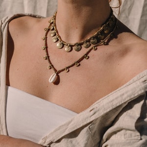 Puede incluir: Dos collares dorados superpuestos. El collar superior presenta colgantes en forma de espiral, mientras que el inferior tiene peque&ntilde;as cuentas y un colgante de concha. Los collares se llevan sobre un top blanco y una camisa beige.