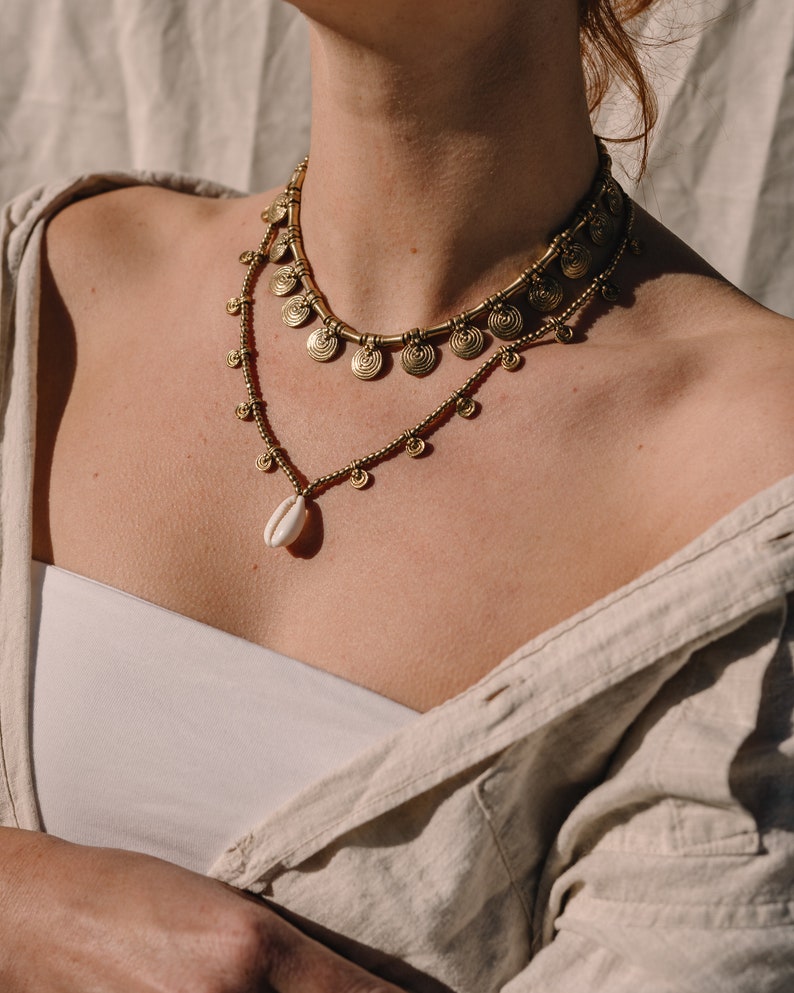 Puede incluir: Dos collares dorados superpuestos. El collar superior presenta colgantes en forma de espiral, mientras que el inferior tiene peque&ntilde;as cuentas y un colgante de concha. Los collares se llevan sobre un top blanco y una camisa beige.