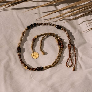 Op de afbeelding: Een bruine en gouden kralenketting met een gouden muntbedel en een bruine koordsliert. De ketting ligt op een witte stoffen achtergrond.