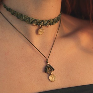 May include: A green macrame choker necklace with a gold pendant featuring a spiral design. The necklace is layered over a simple string necklace with a similar pendant.