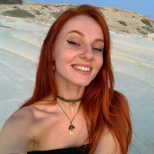 May include: A woman with long, bright red hair is smiling. She is wearing a green and gold choker necklace with a pendant. She is wearing a black top and has a nose ring.