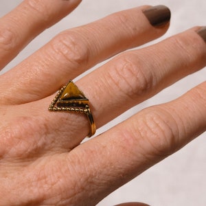 Op de afbeelding: Een gouden ring met een driehoekige tijgeroog-edelsteen. De ring heeft een parelmoerontwerp rond de steen.