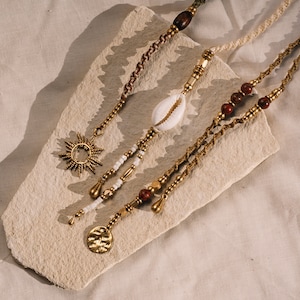 Op de afbeelding: Vier gelaagde kettingen met gouden kralen, bedels en een schelp hanger. De kettingen liggen op een lichtbruin stenen oppervlak.