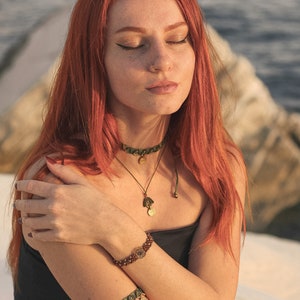 May include: A woman with long, red hair wearing a black top and a green and gold beaded necklace and bracelet. She is wearing a nose ring and has her eyes closed.