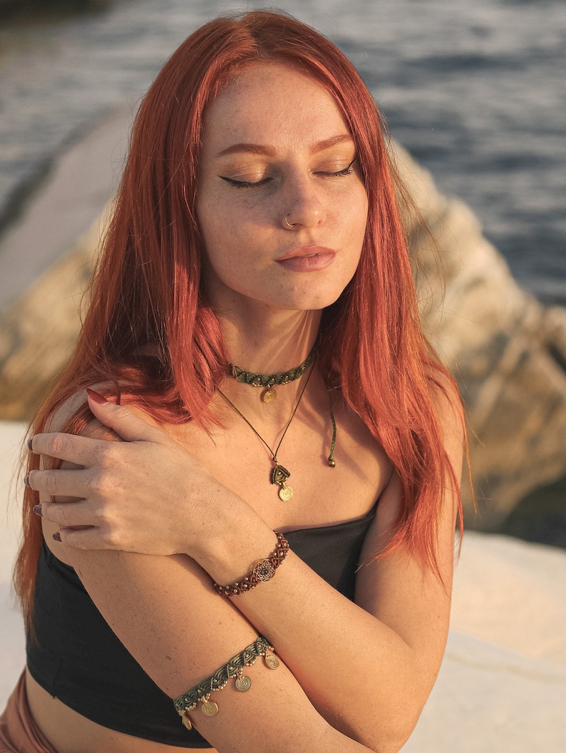 May include: A woman with long, red hair wearing a black top and a green and gold beaded necklace and bracelet. She is wearing a nose ring and has her eyes closed.
