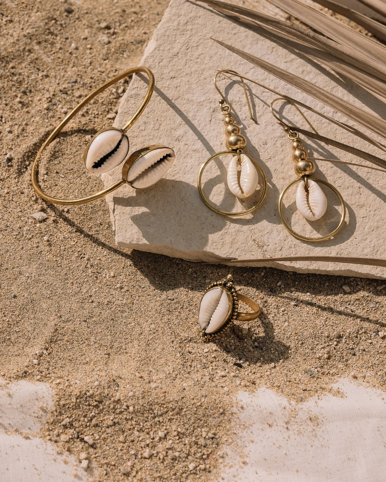 Puede incluir: Un conjunto de joyas doradas con conchas de cauri. El conjunto incluye una pulsera de pu&ntilde;o con dos conchas, un par de pendientes de aro y un anillo. Las joyas se exhiben sobre una superficie arenosa con un fondo neutro.