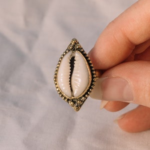 Puede incluir: Anillo dorado con un centro de concha blanca. La concha est&aacute; engastada en un marco decorativo en forma de l&aacute;grima con peque&ntilde;os detalles de cuentas. El anillo se sostiene sobre un fondo blanco suave.