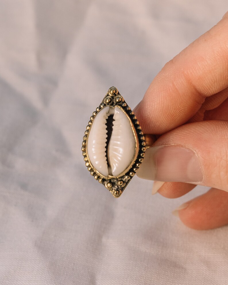 Puede incluir: Anillo dorado con un centro de concha blanca. La concha est&aacute; engastada en un marco decorativo en forma de l&aacute;grima con peque&ntilde;os detalles de cuentas. El anillo se sostiene sobre un fondo blanco suave.