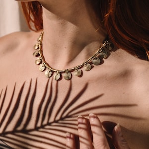 Può includere: Una collana color oro con dettagli a ciondolo a spirale. La collana è indossata attorno al collo. L'immagine mostra un primo piano della collana e della parte superiore del petto. L'ombra di una foglia di palma è proiettata sulla pelle.