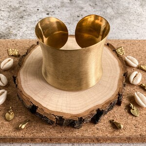 K&ouml;nnte beinhalten: Ein breites goldenes Metall-Armband mit geb&uuml;rsteter Oberfl&auml;che. Das Armband wird auf einer Holzscheibe pr&auml;sentiert, die von kleinen wei&szlig;en Muscheln und goldenen Akzenten umgeben ist.
