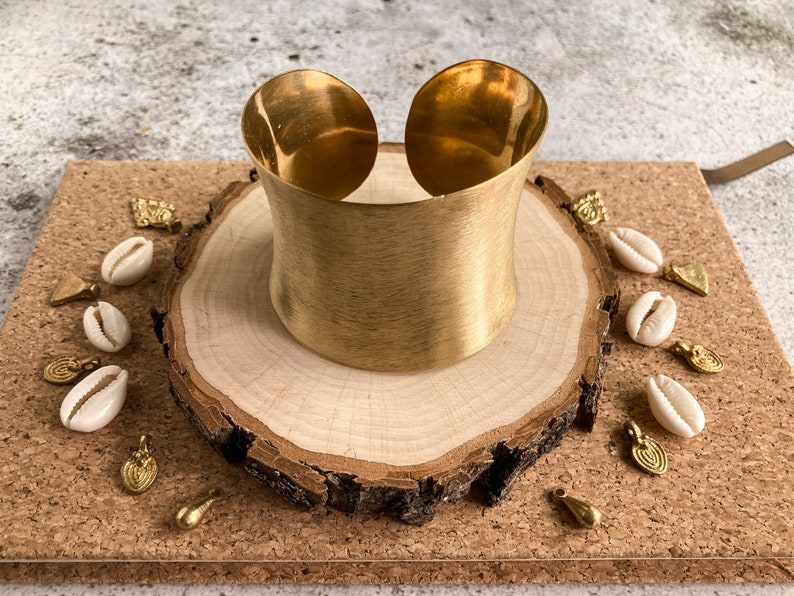K&ouml;nnte beinhalten: Ein breites goldenes Metall-Armband mit geb&uuml;rsteter Oberfl&auml;che. Das Armband wird auf einer Holzscheibe pr&auml;sentiert, die von kleinen wei&szlig;en Muscheln und goldenen Akzenten umgeben ist.