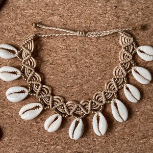 Puede incluir: Un tobillo de macramé beige con conchas de cauri blancas. El tobillo está hecho con un cordón anudado y presenta una variedad de tamaños de conchas.