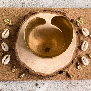 K&ouml;nnte beinhalten: Eine goldene Schale mit einer einzigartigen Form sitzt auf einer Holzscheibe. Die Schale ist von kleinen wei&szlig;en Muscheln und goldenen Anh&auml;ngern umgeben.