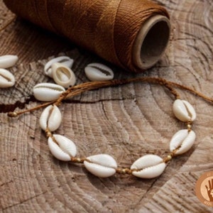 Op de afbeelding: Een bruin koordarmband met witte kauri schelpen en gouden kralen. De armband ligt op een houten oppervlak met een spoel bruin koord op de achtergrond.