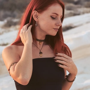 May include: A woman with long red hair wearing a black strapless top, a gold necklace with a pendant, and a gold bracelet on her left wrist. She is also wearing a gold choker necklace and earrings.