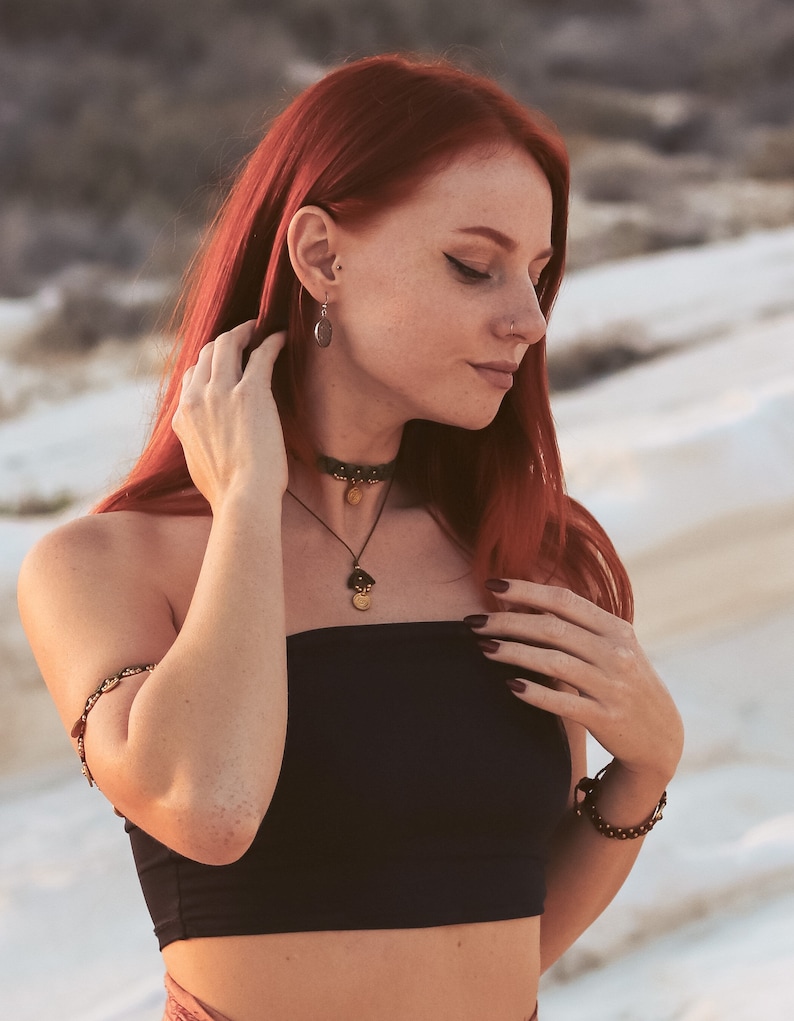 May include: A woman with long red hair wearing a black strapless top, a gold necklace with a pendant, and a gold bracelet on her left wrist. She is also wearing a gold choker necklace and earrings.