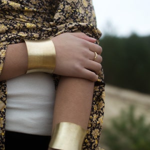 K&ouml;nnte beinhalten: Ein Frauenarm mit zwei goldenen Armreifen. Der Armreif am Unterarm ist breiter als der Armreif am Oberarm. Die Frau tr&auml;gt ein Oberteil mit schwarzem und goldenem Muster.