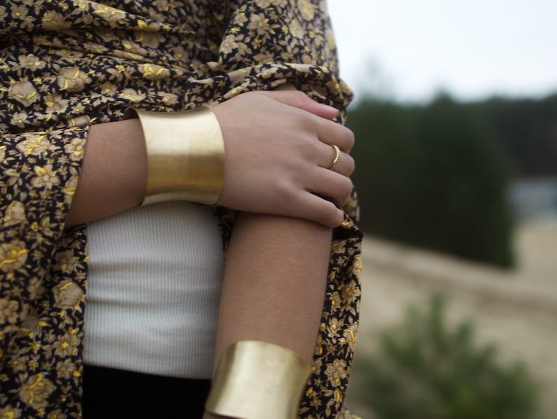 K&ouml;nnte beinhalten: Ein Frauenarm mit zwei goldenen Armreifen. Der Armreif am Unterarm ist breiter als der Armreif am Oberarm. Die Frau tr&auml;gt ein Oberteil mit schwarzem und goldenem Muster.